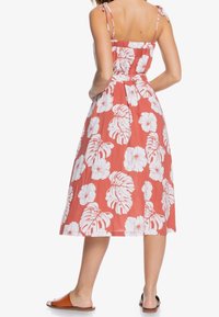 Mujer con un vestido midi sin mangas color coral con estampados de hojas y flores tropicales en blanco, combinado con sandalias planas marrones.