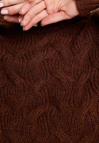 Brown knitted sweater featuring a wavy cable pattern, with soft texture and ribbed cuffs. Close-up of hands resting on the fabric.