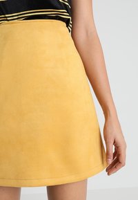 Žlutá semišová sukně s hladkou texturou, rozšířeného střihu a lemem mírně se zvedajícím nahoru. Doprovázená černým pruhovaným topem.