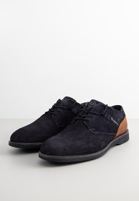 Chaussures en maille bleu foncé avec des accents en daim et un détail en cuir marron sur le talon. Présente un système de laçage et une semelle en caoutchouc plate.