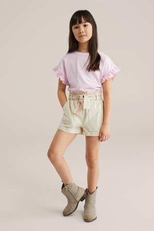 Djevojka stoji s rukama u džepovima, nosi ružičastu majicu kratkih rukava s volanima, bež visokokrojene kratke hlače s remenom i svijetlosmeđe čizme.