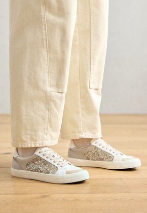 Sneakers w połączeniu białych i beżowych materiałów, z błyszczącym akcentem na boku i teksturowaną podeszwą.