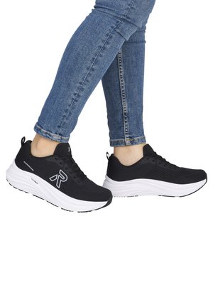 Fødder iført sorte og hvide sports-sneakers med "R" logo, parret med blå tætsiddende jeans mod en hvid baggrund.