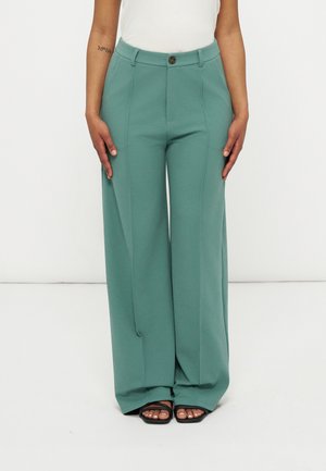 Kobieta ubrana w szerokie spodnie o wysokim stanie w kolorze zielonym morskim z guzikiem i suwakiem z przodu, zestawione z czarnymi sandałami na otwartym palcu i białym topem.