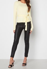 Pull jaune côtelé à manches longues avec une taille nouée, associé à un pantalon skinny noir en similicuir et des sandales à talons ouvertes noires.