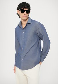 Koszula zapinana na guziki z jasnoniebieskiego materiału chambray; długie rękawy, szpiczasty kołnierz, jedna kieszeń na piersi z haftowanym detalem, noszona z białymi spodniami.