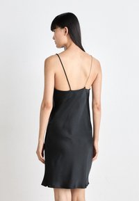 Black satin slip dress with thin straps and a low back. Features soft texture and a knee-length hem with a subtle scalloped edge.
