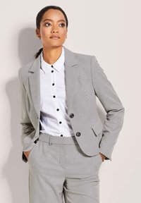 Grau strukturiertes Anzug mit einem maßgeschneiderten Blazer und passenden Hosen. Darunter ein weißes Hemd mit schwarzen Knöpfen. Einfaches Design, schlanke Proportionen.