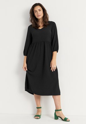 Zwarte jurk met een gathered taille, lange pofmouwen en een V-hals. Combinatie met groene leren hoge hakken. Gladde, gestructureerde stof.