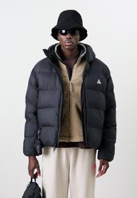 Svart puffjacka med kviltat mönster, brun fleecejacka med dragkedja under, beige byxor, svart bucket hat och solglasögon. Avtagbar väska på handleden.