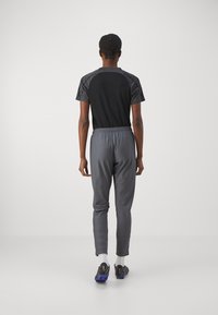 Schwarzes Sportshirt mit strukturierten grauen Ärmeln, kombiniert mit grauen, eng zulaufenden Jogginghosen und schwarzen Sportschuhen mit lila Sohle.
