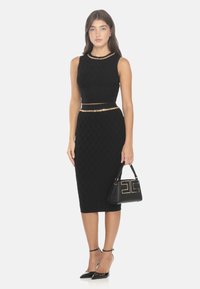 Top corto nero e gonna midi aderente con motivo a rombi testurizzati, accentuati da una cintura in catena dorata. Borsa nera con logo, tacchi a punta.