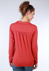 Long-sleeve shirt in coral fabric with a collar, featuring a pleated back design and a slightly curved hem. Worn with blue jeans.