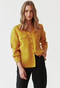 Geel, lange mouwen overhemd met knopen, twee borstzakken, zachte textuur en een V-hals. Draagt met zwarte broek.