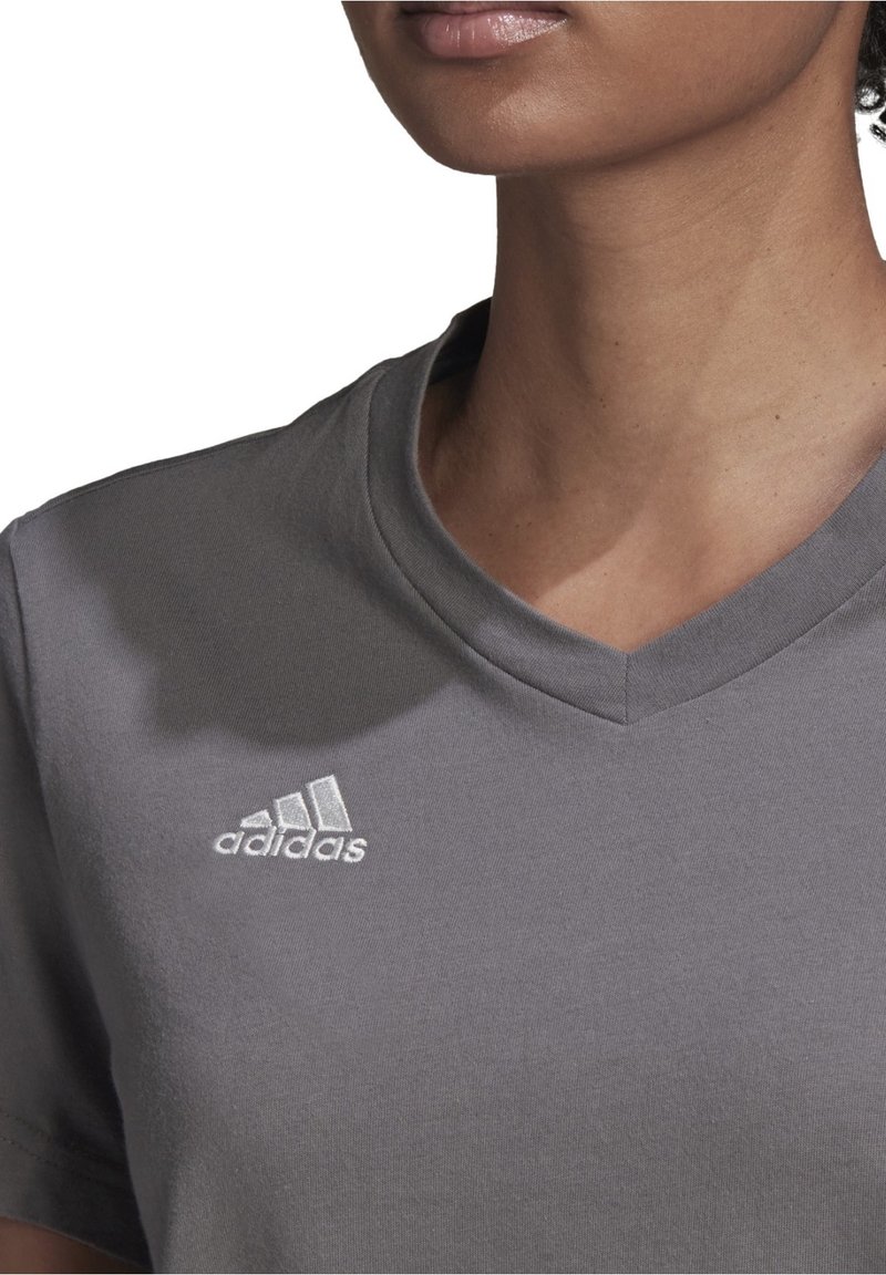 Gros plan sur une personne portant un T-shirt Adidas gris à col en V avec le logo Adidas blanc sur le côté gauche de la poitrine.