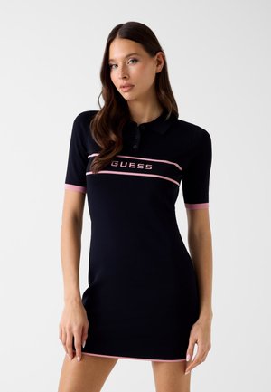 Kobieta o długich brązowych włosach, ubrana w czarną sukienkę polo z krótkim rękawem, różowymi lamówkami i logo „GUESS” na piersi.