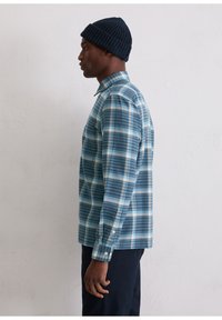 Koszula w kratę w odcieniach niebieskiego i białego, z klasycznym kołnierzykiem i długimi rękawami. Noszona z ciemną, dzianą czapką.
