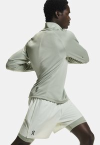 Hellgrünes, langärmliges Sportoberteil aus strukturiertem Stoff, kombiniert mit cremefarbenen Shorts. Beide Kleidungsstücke haben ein Logodetail am Saum.