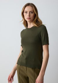 Olivgrünes Kurzarm-Strickoberteil mit rundem Ausschnitt, gerippten Bündchen und Saum. Glatte Textur, taillierte Passform, kombiniert mit passenden Hosen.