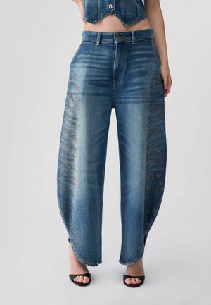 Donna che indossa jeans blu ampi con gamba curva e borchie sui lati, abbinati a sandali con tacco aperto e un top di jeans corto.