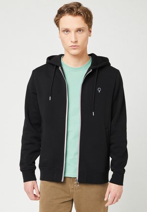 Schwarzer Reißverschluss-Hoodie mit einer Kordelzugkapuze, mit einem dezenten Baum-Logo. Getragen über einem hellgrünen Rundhals-T-Shirt und beigen Hosen.