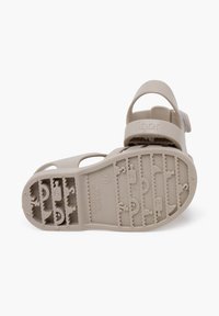 Sandalia infantil beige con suela de goma texturizada, correa de talón ajustable y diseño con perforaciones para mayor transpirabilidad. Presenta un moldeado del logo.