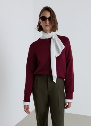 Donna con occhiali da sole scuri, maglione bordeaux, sciarpa bianca annodata al collo, polsini bianchi e pantaloni verde oliva, in piedi davanti a uno sfondo semplice.