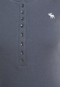 Ribbade, mörkblå henley-tröja med en knappslå med sex mörka knappar och en vit älglogotyp på bröstet. Mjuk bomullstextur.