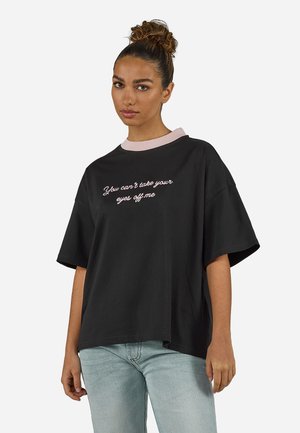 Femeie cu păr creț prins în coc, purtând un tricou negru larg cu guler roz și textul „Nu-ți poți lua ochii de la mine” și blugi albastru deschis.