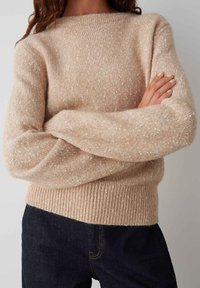 Pull en tricot beige avec un motif texturé, un ourlet côtelé et des manches larges. Associé à un jean en denim foncé.
