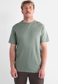 T-shirt en coton vert olive avec un col rond, des manches courtes et un petit logo clair sur la poitrine. Design simple, coupe décontractée.