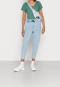 Ljusblå denimgujer, grön ribbat kortärmad topp, vita sportiga sneakers och en grå mönstrad axelväska med logotyp.