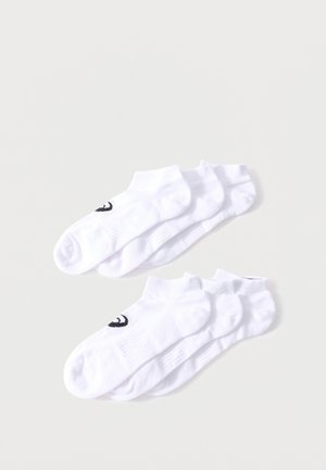 Dos juegos de tres calcetines blancos hasta el tobillo con logotipos en forma de media luna negros apilados sobre un fondo gris claro.