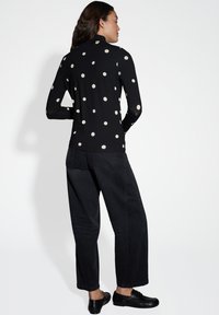 Zwarte longsleeve top met grote crèmekleurige stippen, gecombineerd met brede zwarte jeans en zwarte schoenen, bekeken vanuit de achterkant.