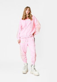 Sudadera rosa y pantalones jogger a juego con bordados de plumas en las mangas y las piernas. Incluye cordones y bolsillos laterales. Combinado con botas blancas.