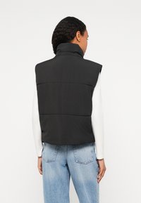 Μαύρο επενδυμένο γιλέκο με ψηλό γιακά, με κουβαρδιστή υφή και κομμένο σχέδιο, φορεμένο πάνω από ένα άσπρο π�ουκάμισο με μακριά μανίκια και μπλε τζιν.