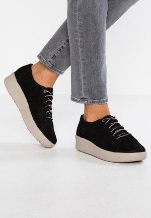 Czarne zamszowe sneakersy z beżową gumową podeszwą, z metalicznymi sznurówkami i logo na boku. Design ma niski profil i zaokrąglony czubek.