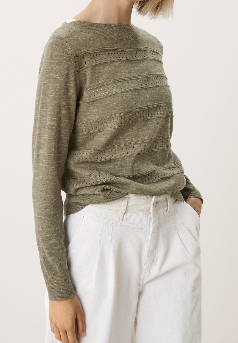 Pull tricoté vert olive avec rayures horizontales et motifs ajourés, assorti à un pantalon blanc taille haute. Texture lisse, encolure ronde.