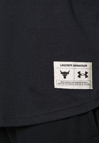 Czarna bawełniana koszulka z krótkim rękawem z boczną metką z grafiką byka, logo Under Armour oraz tekstem opisującym odzież sportową.