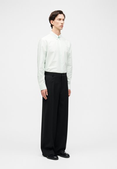 Άνδρας όρθιος, ντυμένος με λευκό πουκάμισο με κουμπιά μέσα σε μαύρο παντελόνι φαρδύ στο πόδι, με μαύρα επίσημα παπούτσια, ουδέτερο φόντο.