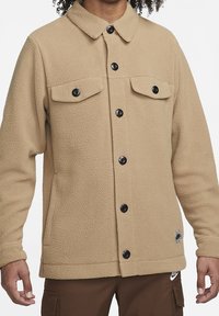 Beżowa kurtka polarowa z przodem zapinanym na guziki, dwoma kieszeniami na piersi i czarnymi guzikami jako akcentami. Posiada zaokrąglony kołnierz oraz teksturowaną tkaninę.