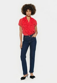 Rote Bluse mit Rüschendetails, kurzen Ärmeln und Kragen, kombiniert mit dunkelblauen, taillierten Jeans und schwarzen, hohen Mules.
