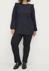 Granatowa tunika z falbaniastymi akcentami, długimi rękawami, teksturowana tkanina, zestawiona z czarnymi dopasowanymi spodniami i czarnymi butami slip-on.