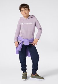 Un jeune garçon portant un hoodie lavande de Champion, un pantalon de jogging marine et des baskets vert olive, se tenant avec un sweat-shirt lavande noué autour de la taille.