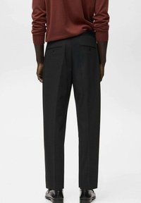 Osoba nosząca czarne dopasowane spodnie, brązowy długi rękaw i czarne eleganckie buty, pokazana z tyłu na jednolitym tle.