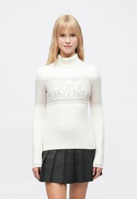 Hvit turtleneck-genser med lysegrå horisontale striper og logoen "Le Ger Ski Club". Har lange ermer og ribbestrikkede mansjetter. Svart plissert skjørt.