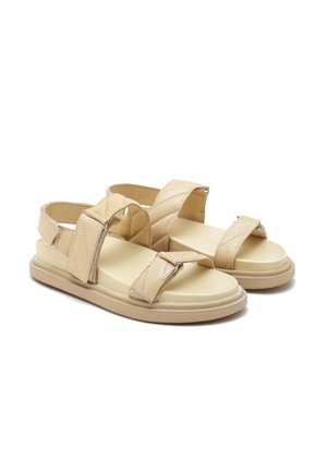 Beige sandalen met stoffen kruisbanden en een verhoogde zool. Het zachte materiaal heeft zichtbare stiksels en een platte, brede voetbed.