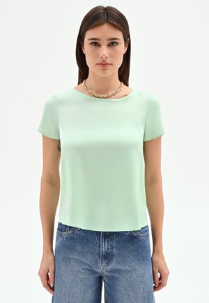 Femme portant un haut à manches courtes vert clair, un jean taille haute bleu et un collier doré épais, debout devant un fond uni.