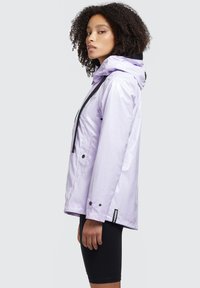 Jachetă waterproof de culoare violet deschis, cu glugă, accente negre și șnururi ajustabile. Dispune de butoane tip snap și are o croială relaxată.