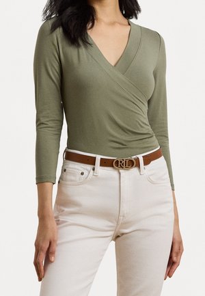 Vrouw draagt een olijfgroene, lange mouwen wikkeltop, ingestopt in een witte, hooggetailleerde jeans met een bruine riem met een gouden RL-gesp.